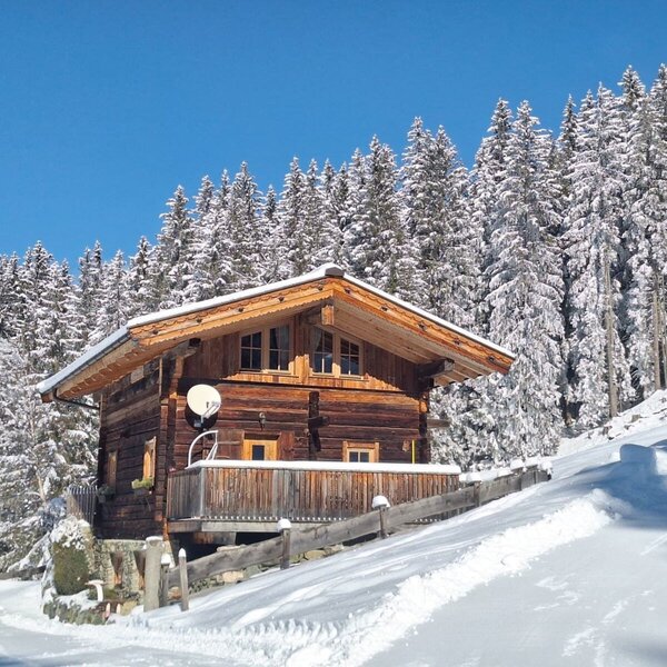 Die Alm als Holzhaus im Winter, umgeben von tief verschneiten Wäldern und mit einer kleinen Terrasse.