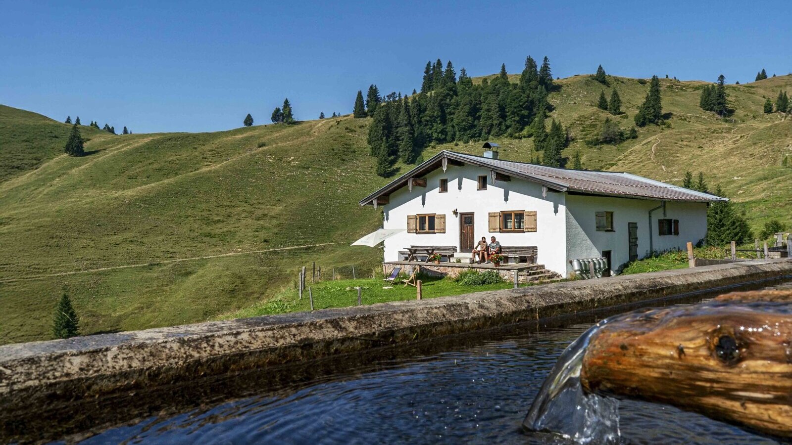 Die Alm mit ihrer Terrasse und einem Wasserbrunnen im Vordergrund, eingebettet in eine grüne Berglandschaft.