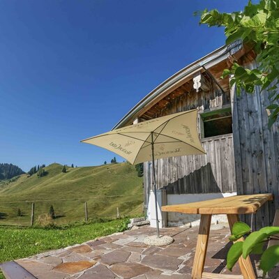Die Terrasse der Alm mit einem Holztisch, einer Bank, einem Sonnenschirm und Blick auf die grünen Hügel.
