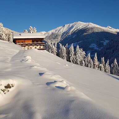 Der Bauernhof, umgeben von einer tief verschneiten Berglandschaft und Tannenbäumen.