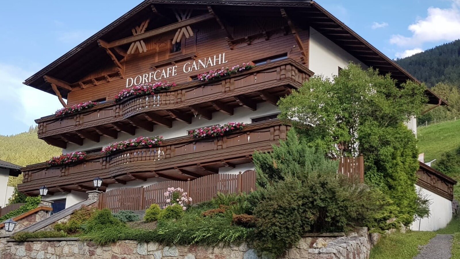 Die Außenansicht des Bauernhofs mit traditionellen Holzbalkonen, die mit Blumen geschmückt sind, und einer Steinmauer.