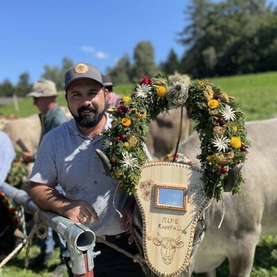 Der Bauer mit einer traditionell geschmückten Kuh, die einen Blumenkranz und eine Holztafel trägt, auf dem Bauernhof.