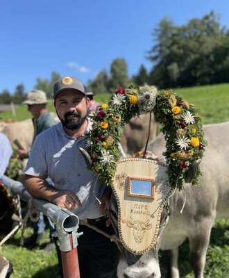 Der Bauer mit einer traditionell geschmückten Kuh, die einen Blumenkranz und eine Holztafel trägt, auf dem Bauernhof.
