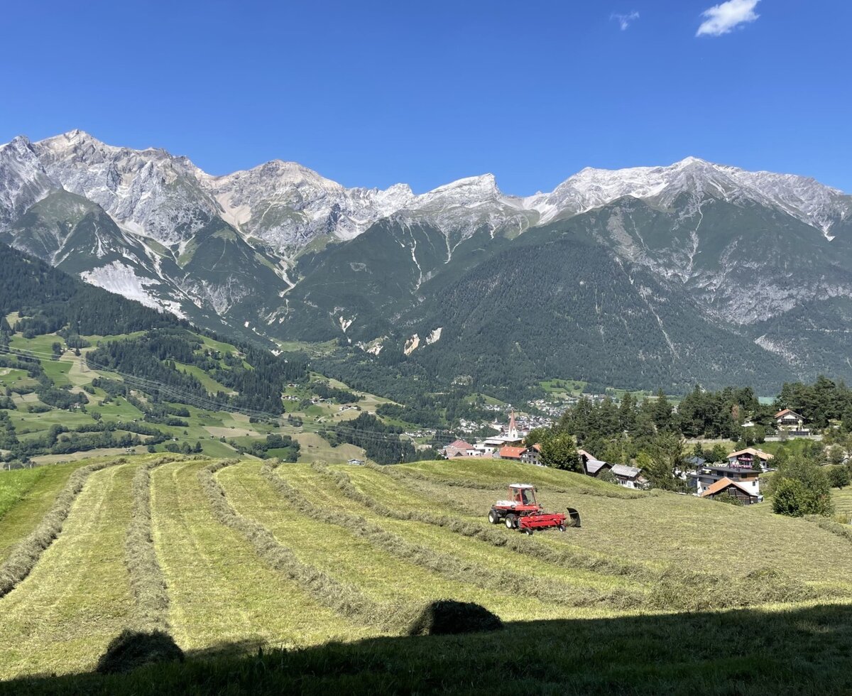 Ein gemähtes Feld des Bauernhofs mit Traktor, Bergpanorama und einem Dorf im Hintergrund.