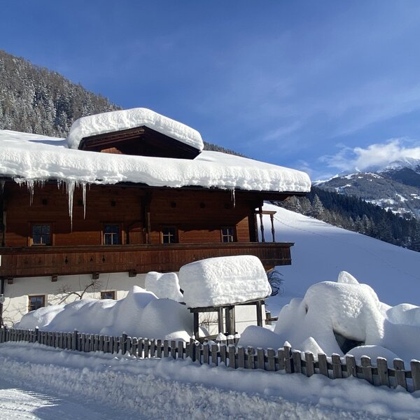 Der Bauernhof im Winter mit schneebedecktem Dach, Eiszapfen und den umliegenden Bergen.