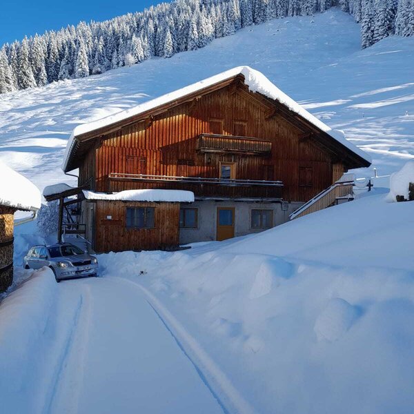 Die Alm als Holzgebäude im Winter, umgeben von verschneiten Bergen und Wald, mit einer geräumten Zufahrt.