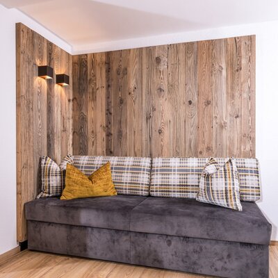 Sitzbereich im Bauernhof mit einem dunkelgrauen Sofa, karierten Kissen, Holzwand und Wandleuchten.
