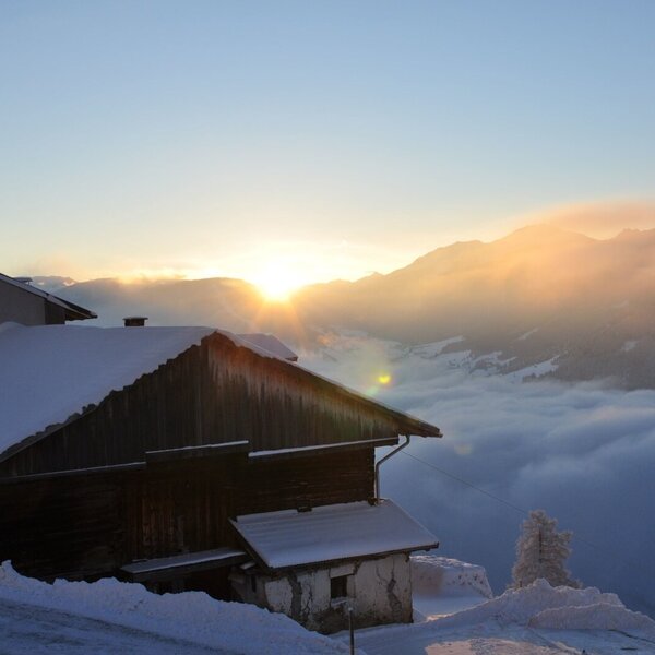 Die winterliche Umgebung des Bauernhofs mit schneebedeckten Gebäuden, Bergen und Talnebel bei Sonnenaufgang.