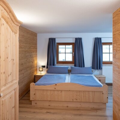 Schlafzimmer im Ap­par­te­ments mit Holz-Doppelbett, Holzschrank und zwei Fenstern.