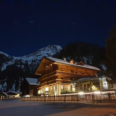 Außenansicht des beleuchteten Bauernhofs bei Nacht, umgeben von Schnee und verschneiten Bergen.