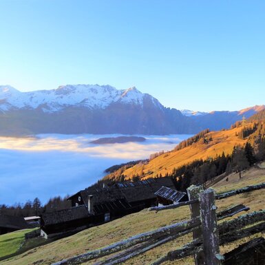 Die Alm mit ihren Gebäuden und einem Holzzaun im Vordergrund bietet einen Blick auf schneebedeckte Berge und Nebel im Tal.