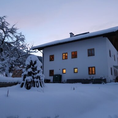 Der Bauernhof im Winter, umgeben von Schnee und mit beleuchteten Fenstern.