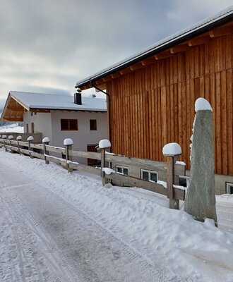 Der Bauernhof im Winter mit verschneitem Weg und Holzzaun vor den Gebäuden.