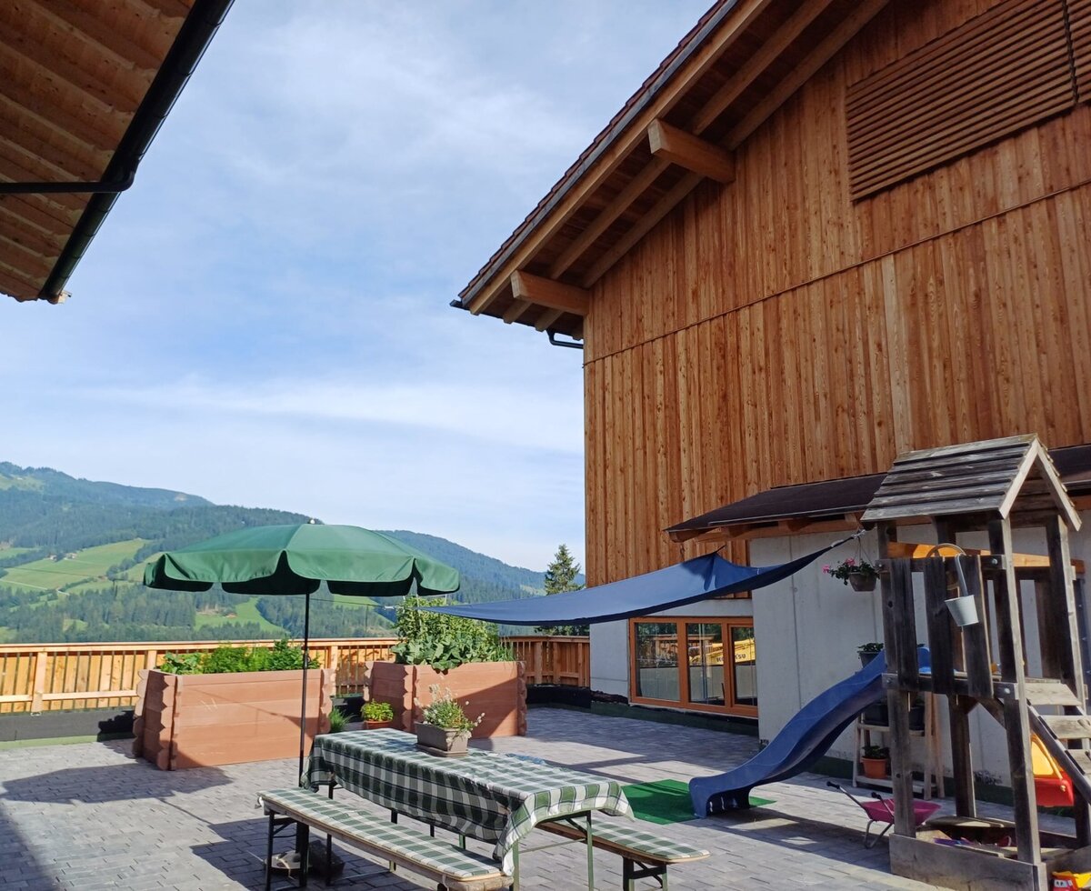 Außenterrasse des Bauernhofs mit Esstisch, Sonnenschirm, Kinderspielplatz und Bergblick.