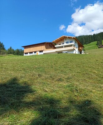 Der Bauernhof mit modernem Wohnteil und angrenzendem Wirtschaftsgebäude, gelegen auf einer grünen Wiese am Hang.