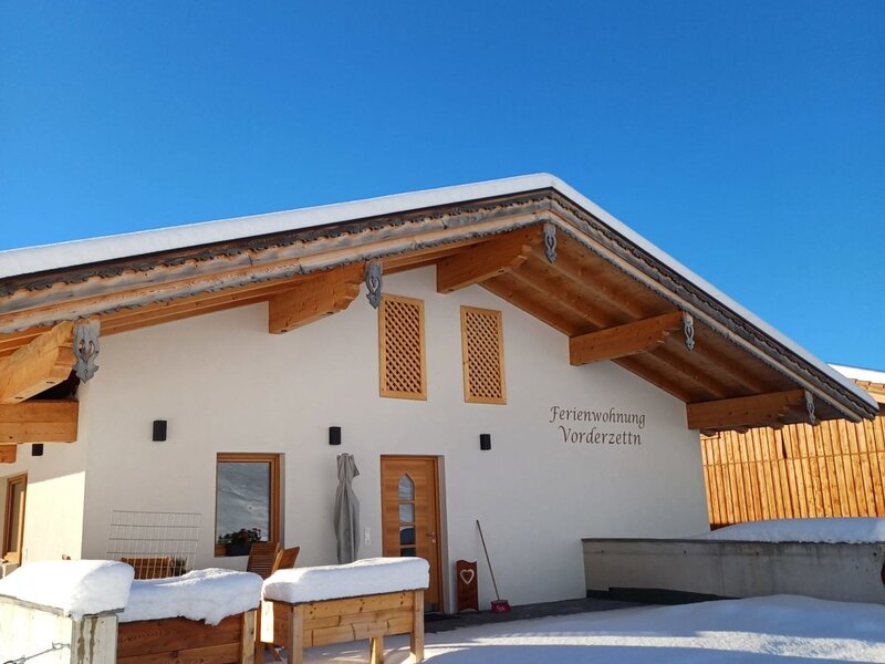 Die winterliche Außenansicht des Bauernhofs 'Ferienwohnung Vorderzettn', bedeckt mit Schnee unter blauem Himmel.