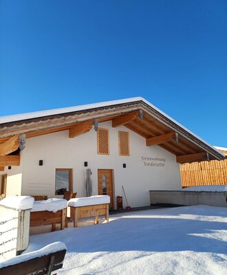 Die winterliche Außenansicht des Bauernhofs 'Ferienwohnung Vorderzettn', bedeckt mit Schnee unter blauem Himmel.