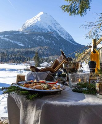 Ein liebevoll gedeckter Außentisch des Appartements mit herzhaften Snacks vor einer malerischen, verschneiten Bergkulisse.