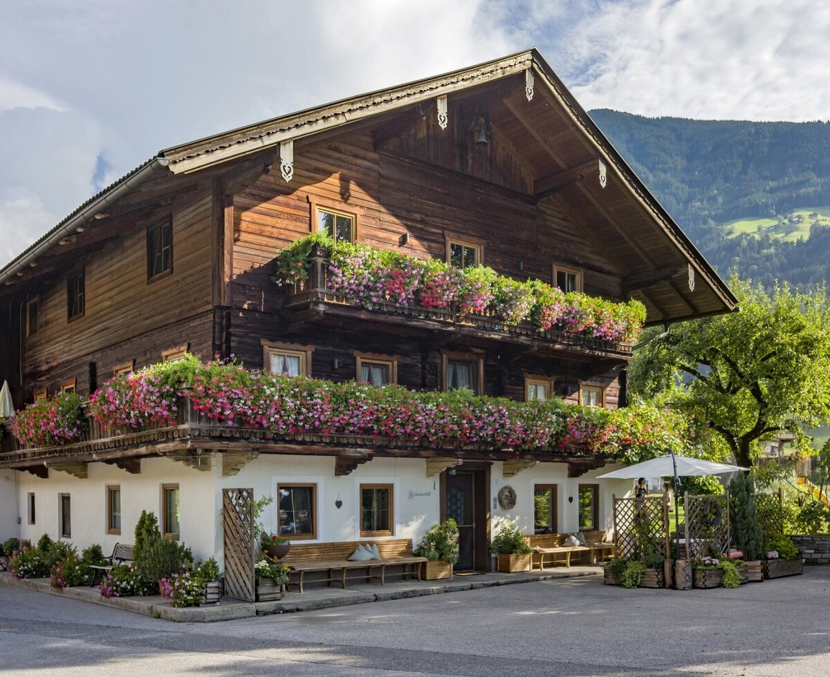 Der Bauernhof Schustererhof mit traditioneller Holzfassade, blumengeschmückten Balkonen und Sitzgelegenheiten im Außenbereich vor Bergkulisse.