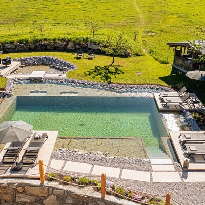 Der Naturschwimmteich des Bauernhofs mit Sonnenliegen, Sonnenschirmen und einer Sitzgruppe im Garten.