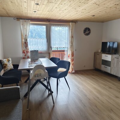Wohn- und Essbereich der Ferienwohnung mit Esstisch, Sitzbank, Stühlen, Kinderhochstuhl und Fernseher, sowie Zugang zum Balkon.