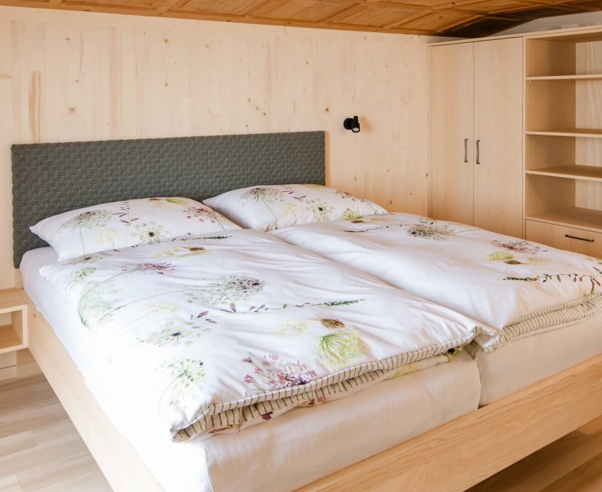 Doppelzimmer im Bauernhof mit Holzwänden und -decke, floraler Bettwäsche, Nachtkästchen mit Leselampen und einem Kleiderschrank mit offenen Regalen.