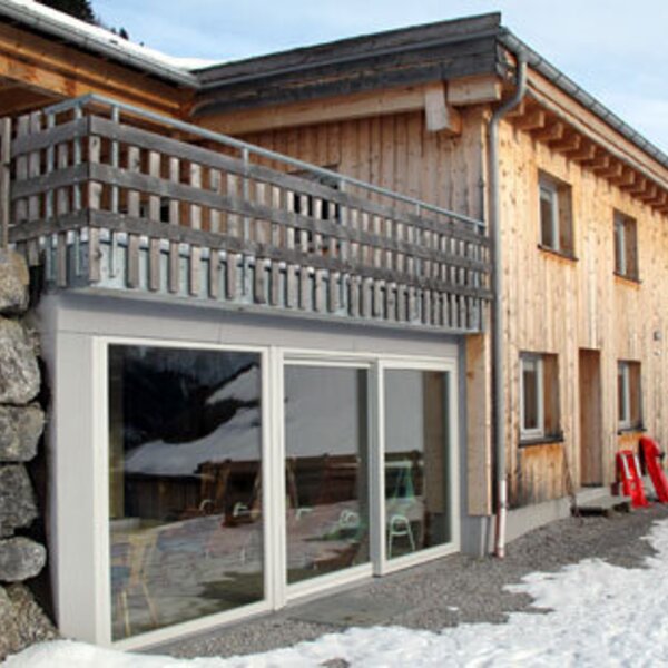 Die Außenansicht des Bauernhofs mit Holzfassade, Balkon, großen Fenstern im Erdgeschoss und einer Natursteinmauer, umgeben von Schnee.