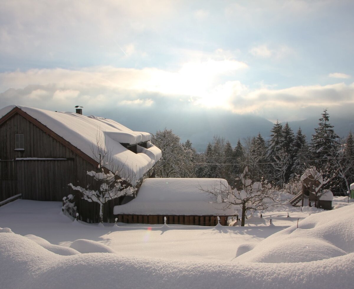 Winterliche Ansicht des Bauernhofs mit verschneiten Gebäuden und einer Bergkulisse unter strahlender Wintersonne.