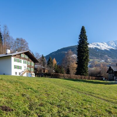 Der Bauernhof mit weißer Fassade und grünen Fensterläden liegt auf einer Wiese mit Blick auf die Berge.