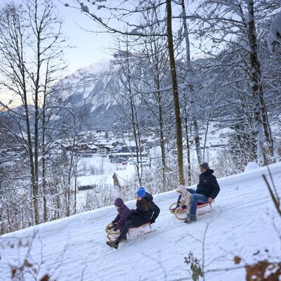Familie beim Rodeln auf einem verschneiten Hang mit Blick auf das Dorf und die Berge, eine Winteraktivität in der Umgebung des Bauernhofs.