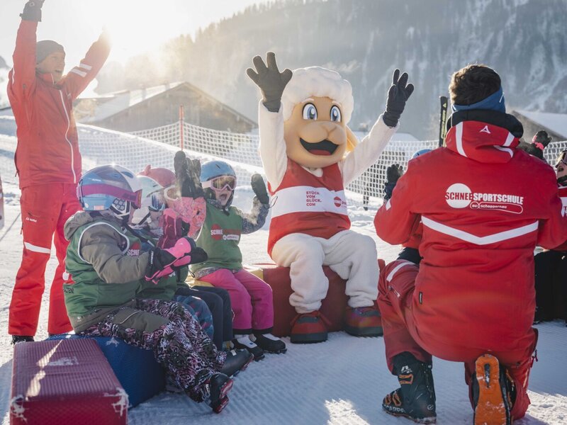 Ein fröhlicher Skikurs für Kinder nahe dem Bauernhof mit einem freundlichen Maskottchen im glitzernden Schnee.