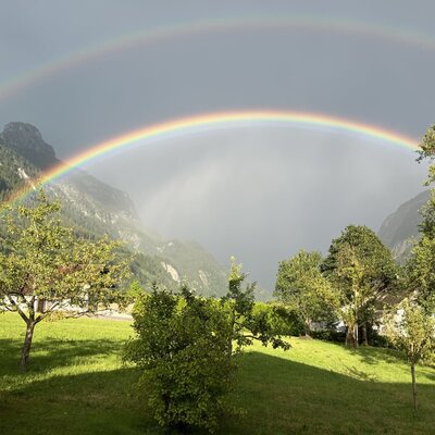Doppelter Regenbogen über den Bergen, sichtbar vom Bauernhof mit grüner Wiese und Bäumen.