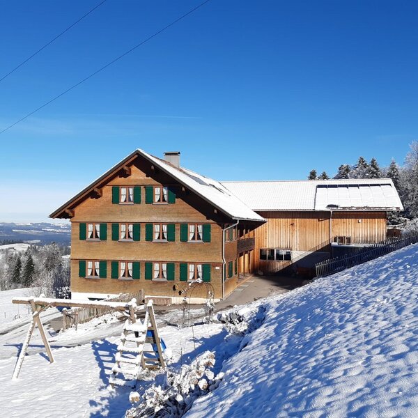 Der Bauernhof im Winter mit schneebedeckten Dächern, umgeben von verschneiter Landschaft und einem Spielgerüst.