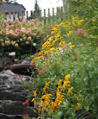 Der Garten des Bauernhofs mit verschiedenen blühenden Pflanzen, einem Natursteinelement und einem Wasserauslauf.