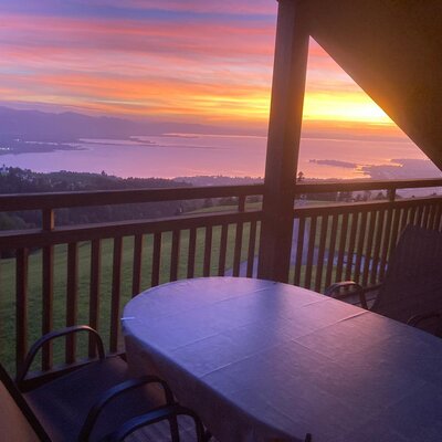 Der einladende Balkon der Ferienwohnung auf dem Bauernhof mit einem Tisch, Stühlen und einem malerischen Ausblick auf den See bei Sonnenuntergang.