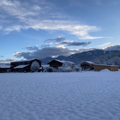 Der Bauernhof in verschneiter Winterlandschaft mit Feldern und Bergen im Hintergrund.
