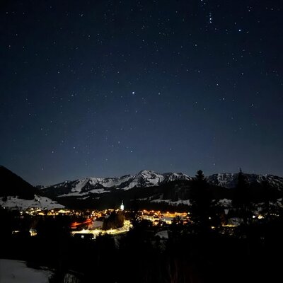 Nachtansicht des Dorfes mit beleuchteten Häusern und einer Kirche, schneebedeckten Bergen und Sternenhimmel, sichtbar vom Bauernhof.