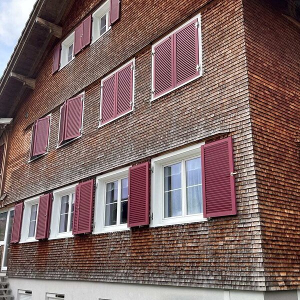 Außenansicht des Bauernhofs mit Holzschindelfassade, weißen Fenstern und roten Fensterläden.