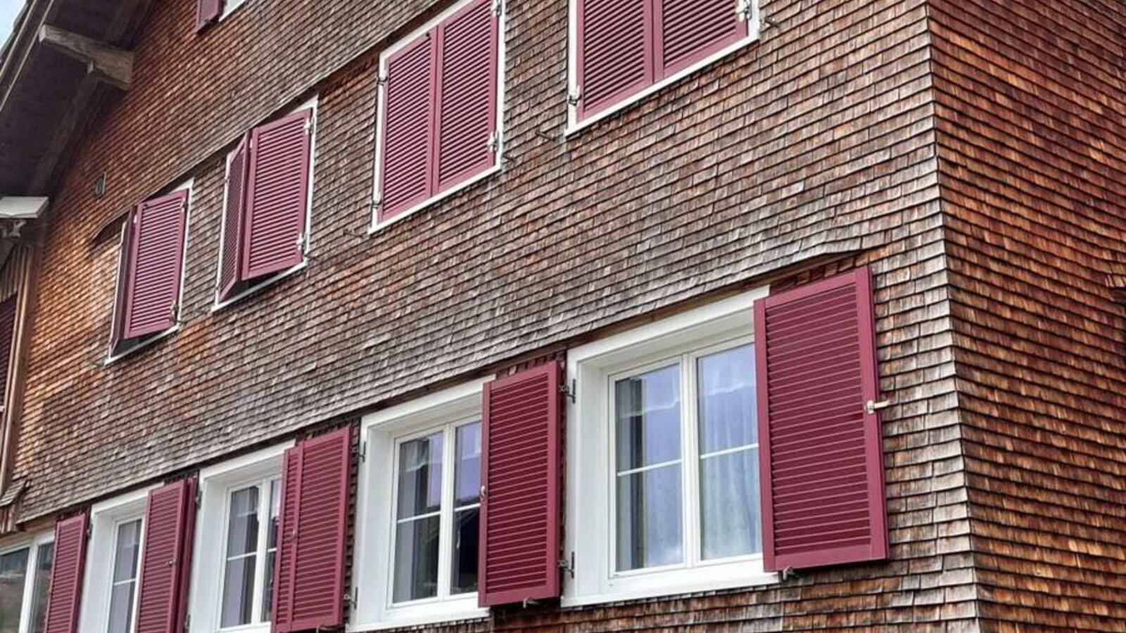 Außenansicht des Bauernhofs mit Holzschindelfassade, weißen Fenstern und roten Fensterläden.