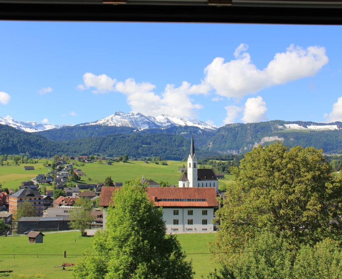 Blick aus dem Fenster auf das Dorf mit Kirche, grüne Felder und schneebedeckte Berge.