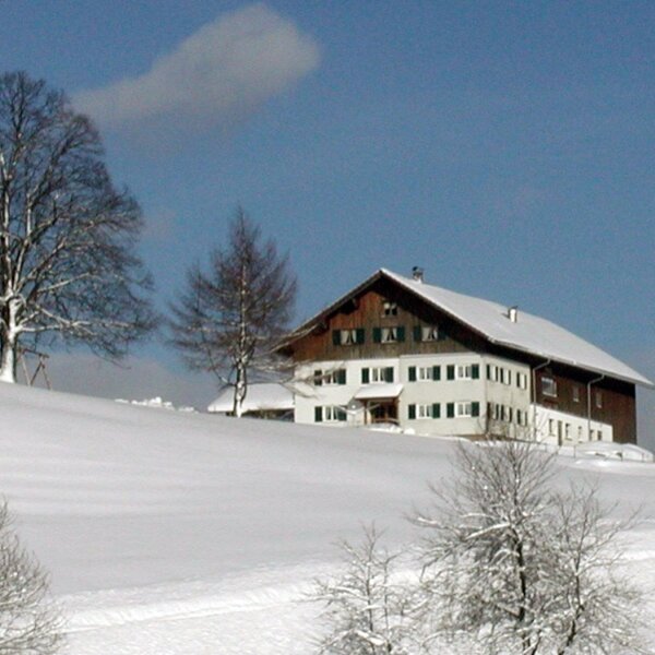 Außenansicht des Bauernhofs mit schneebedecktem Dach und grünen Fensterläden in einer winterlichen Hügellandschaft.