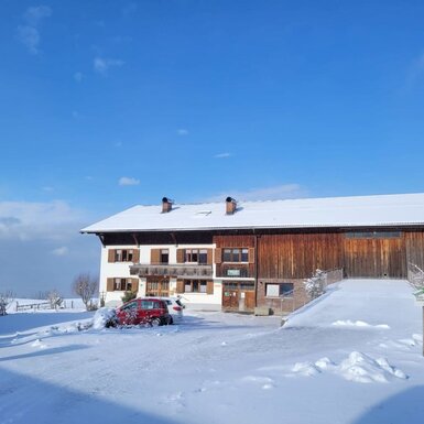 Der Bauernhof im Winter mit schneebedecktem Dach, weißer Fassade und angrenzender Holzscheune unter blauem Himmel.