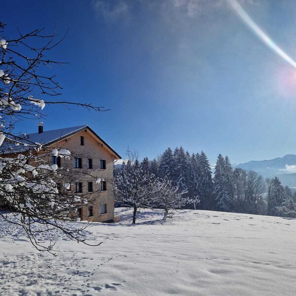 Der Bauernhof in einer verschneiten Winterlandschaft mit Bäumen und Bergen unter blauem Himmel.