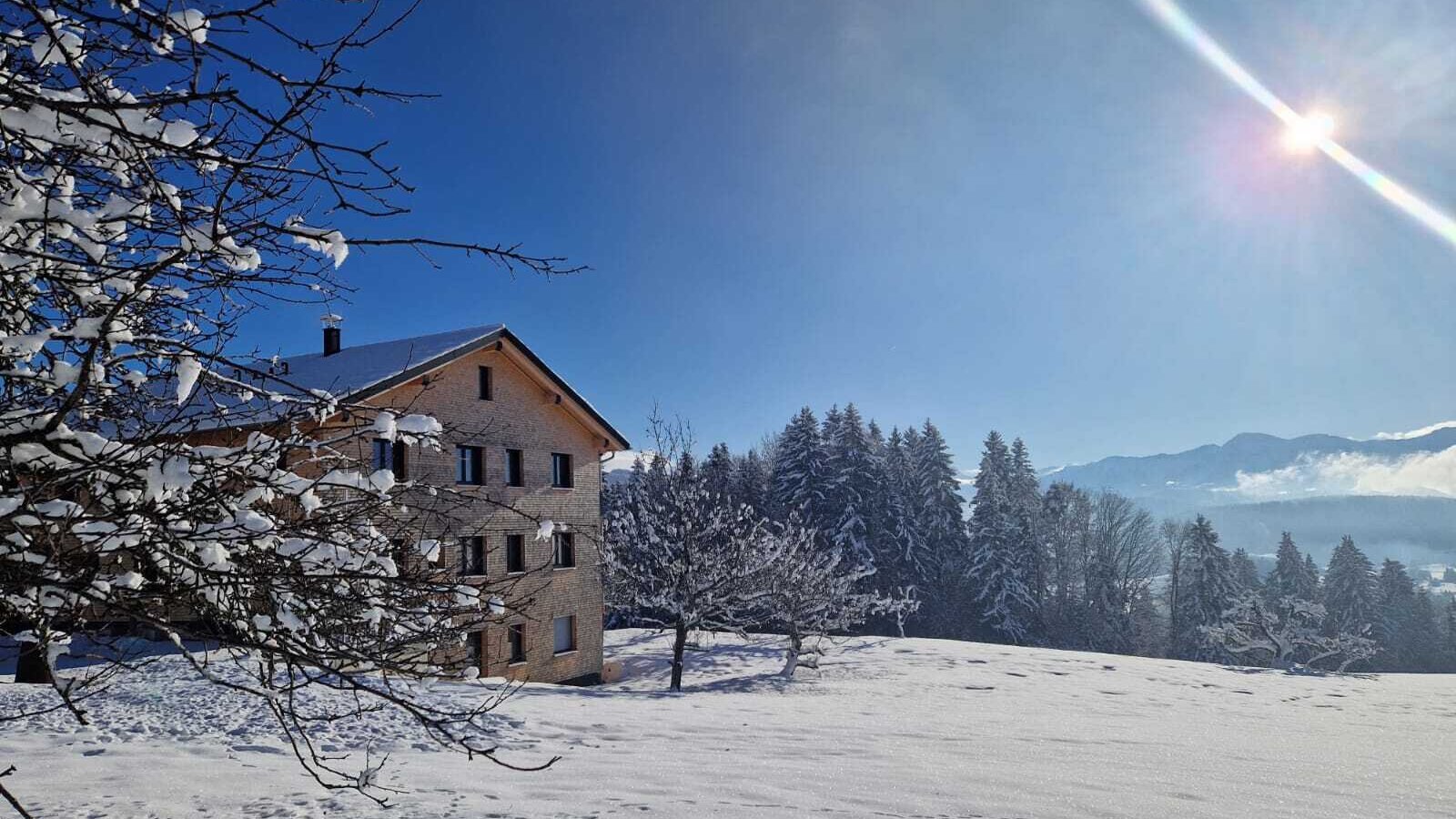 Der Bauernhof in einer verschneiten Winterlandschaft mit Bäumen und Bergen unter blauem Himmel.